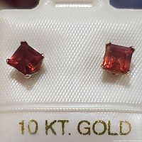 Pair of square cut garnet stud earrings in 10K yellow gold set in a white display with '10 KT. GOLD' marking.