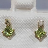 Close-up of the pair of 10K yellow gold earrings with peridot and diamond stones on display stand, showing the green peridot and white diamonds clearly.