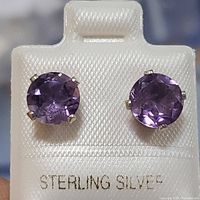 Front view close-up of pair of round cut amethyst earrings set in sterling silver showing purple stones and four-prong setting on textured white display.