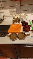 Photo showing the entire lot with vintage Osterizer blender, soup bowls with lids, two ceramic pans, and orange mold on a kitchen counter.