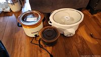 Three Rival crock pots of different sizes photographed on wooden floor: small black/brown, medium beige/brown, large white with controls
