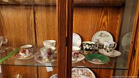 Six assorted cup and saucer sets along with additional saucers and sugar- creamer set displayed on a glass shelf within a wooden cabinet.