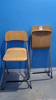 Two wood and metal folding stools against a blue background, showing front and back views.
