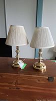 Pair of brass table lamps with white pleated lampshades, placed on wooden surface against a plain wall.