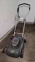 Full view of green electric lawnmower with metal handle and four wheels, placed on concrete floor near wall.