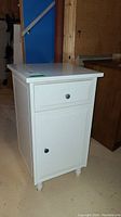 Side view of the white laminate side storage table showing drawer and cabinet door with metal knobs, standing on rounded feet, placed on a concrete floor.