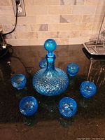 Blue glass decanter with six matching shot glasses arranged around it on a black countertop.