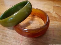 Two round bangle bracelets, one olive green and one amber, shown stacked and side by side on a wood surface.