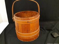 Frontal view of the antique wooden bucket showing the handle, wood grain, and metal bands.