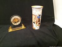 Photo showing the set of two items: the desk clock on a gold-tone base and the tall cylindrical Pickwick vase with decorative paintings.