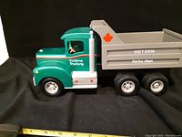 Left-side view of handmade wooden dump truck showing green cab, gray dump bed with red maple leaf decal and white lettering 'Victoria Trucking' and 'Victoria Parks Dept'. Six black and silver wheels visible.