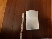 Large silver tone rectangular lighter next to measuring tape showing height approximately 8 inches, placed on wooden floor.