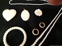 Top view of carved jewelry pieces on black background including heart pendant, cameo pendant, floral brooch, two rings, twisted bangle, and beaded necklace.
