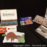 Vintage Grumbacher oil pastels in white box with visible pastels, smaller colorful paint set, decorative storage box, and canvas pouch with sloth design.