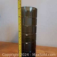 Front view of the tall dark glass vase next to a measuring tape, showing approximately 9.5 inches in height. Segmented horizontal ridges visible on the vase body.