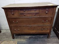Front view showing all six drawers, mixed hardware with round wooden knobs on the upper smaller drawers and metal handles on the larger drawers, medium brown wood with scratches and wear.