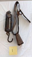 Photo showing the full view of the antique metal hook scale with hanging hook and attached cow bell on leather strap, with yellow paper labeled '1'.