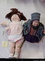 Two dolls lying side by side on a white cloth, showing full body of the Cabbage Patch doll on left and plush old woman doll on right.