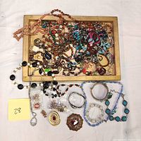 Overview of a mixture of costume jewelry including necklaces, bracelets, brooches and earrings spread over a wooden tray and surrounding area.