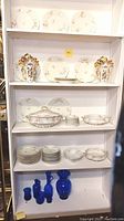 Full shelf view showing white Limoges china plates, bowls, tureen, and cobalt blue glassware at bottom shelf with gilded floral vases on middle shelf.
