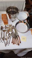 Full view of lot on white tablecloth showing Sango Allure floral bowl, white bowl, metal ice bucket, wooden rolling pin with holder, wooden cutlery block, and array of silver-plated flatware and knives