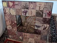 Large square decorative mirror standing against the wall. The frame is covered in square laminate tiles featuring various floral designs in muted purples and pinks.