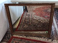 Front view of the rectangular gilt ornate mirror laying on a patterned carpet.