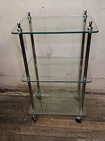 Front angle view showing the three glass shelves, metal poles, and caster wheels on a wooden floor and a beige wall background.