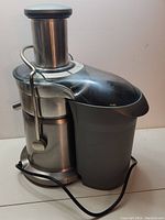 Side view of the Breville Juice Fountain Elite metal juicer showing main body, chute, and juice spout with attached pulp container area (pulp container missing). Power cord visible on surface.