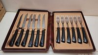 Flatware set open to show 6 knives on left side with stainless steel serrated blades and black handles, and 6 forks on right side with stainless steel tines and black handles inside brown box.