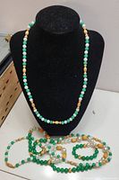 Photo of a green, white, and gold bead necklace on black bust with two additional necklaces lying below it.