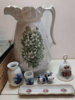 Large ceramic floral pitcher, miniature pitcher and cup, Delft cow figurine, commemorative bell, and rectangular dish shown together