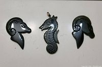 Three dark jade animal pendants on white surface: one seahorse pendant in center with textured body and two ram head pendants with curled horns flanking it
