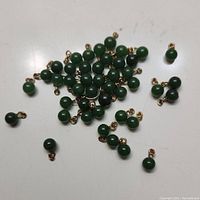 Photo showing scattered collection of small dark green jade beads with attached gold-tone loops on a white background.
