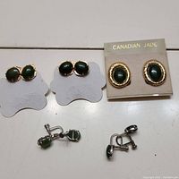 Five pairs of green jade clip-on earrings displayed on table: two pairs on white earring holders, one pair on 'Canadian Jade' card, two pairs loose with clip-on backs visible.
