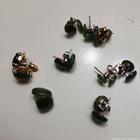 Top-down view of nine pairs of stud jade earrings arranged loosely showing variety in shape and metallic backings.