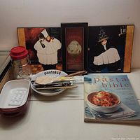 Full lot view showing cookbook, pasta bowl, ceramic dishes, utensils, jar, and wall art