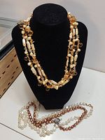Three pearl necklaces displayed on a black velvet bust and laid out beneath it. The multi-strand necklace has irregular cream pearls and light brown crystals. Two single strands of round pearls, white and brownish pink, are also shown.