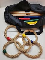 Open carrying bag showing four colored wooden poles (red, blue, yellow, green) and five rope rings with colored tips matching poles.