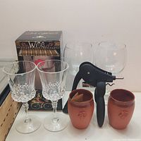Photo showing all items in the lot: lever wine opener, boxed art glass wine glass, two clear crystal cut stemmed wine glasses, two clear plastic wine glasses, and two brown earthenware wine cups.