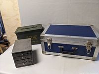 Three cases arranged together on a white surface: green metal military ammo box, blue hardcase with metal edges, small grey metal drawer box.