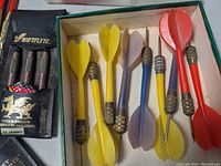 Close-up photo of a set of 6 darts with yellow, purple, and red flights inside a box, showing metal barrels and plastic flights.