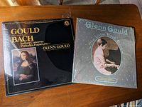 Photo of two Glenn Gould vinyl albums on a wooden table showing the front covers. Left album is black with Mona Lisa image, right album has polka dot cover with Glenn Gould playing piano.