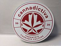 Front view of round metal sign showing Cannadictiva branding and cannabis leaf graphic with red and white color scheme.