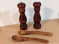 Two tall mid century Barbocraft wooden salt and pepper grinders with a pair of wooden salad utensils (spoon and fork) on a tan surface.