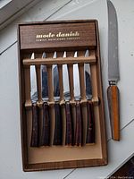 Six serrated steak knives with dark wood handles displayed inside a wooden box labeled 'mode danish FAMILY HEIRLOOMS FOREVER' alongside a larger 13 inch knife with light brown handle.