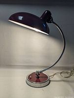 Maroon modernist desk lamp lit up, showing curved chrome arm, round base with toggle switch, and large rounded shade.