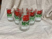Seven cylindrical juice glasses with a rose and picket fence motif displayed upright in a semi-circle.