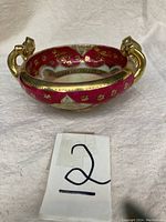 Side angle of the candy dish showing red enamel exterior, gold handles, and gold floral detailing.