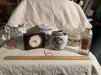 Photo showing 7 vintage items including clock in wooden case, two glass bottles (one labeled Syrup Squill's Dose), two porcelain jars with lids, glass bowl, and blue and white pitcher.